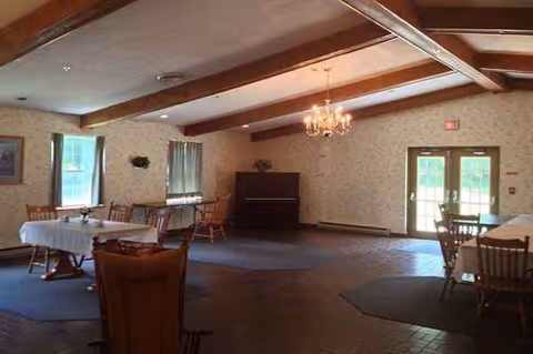 Empty dining room with tables, chairs, chandelier, wood beams, and a piano in a senior home.