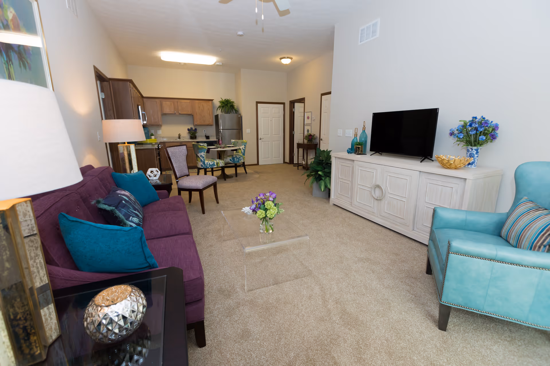 Bright furnished living room with a purple sofa, teal armchair, TV console and an open kitchen in the background.