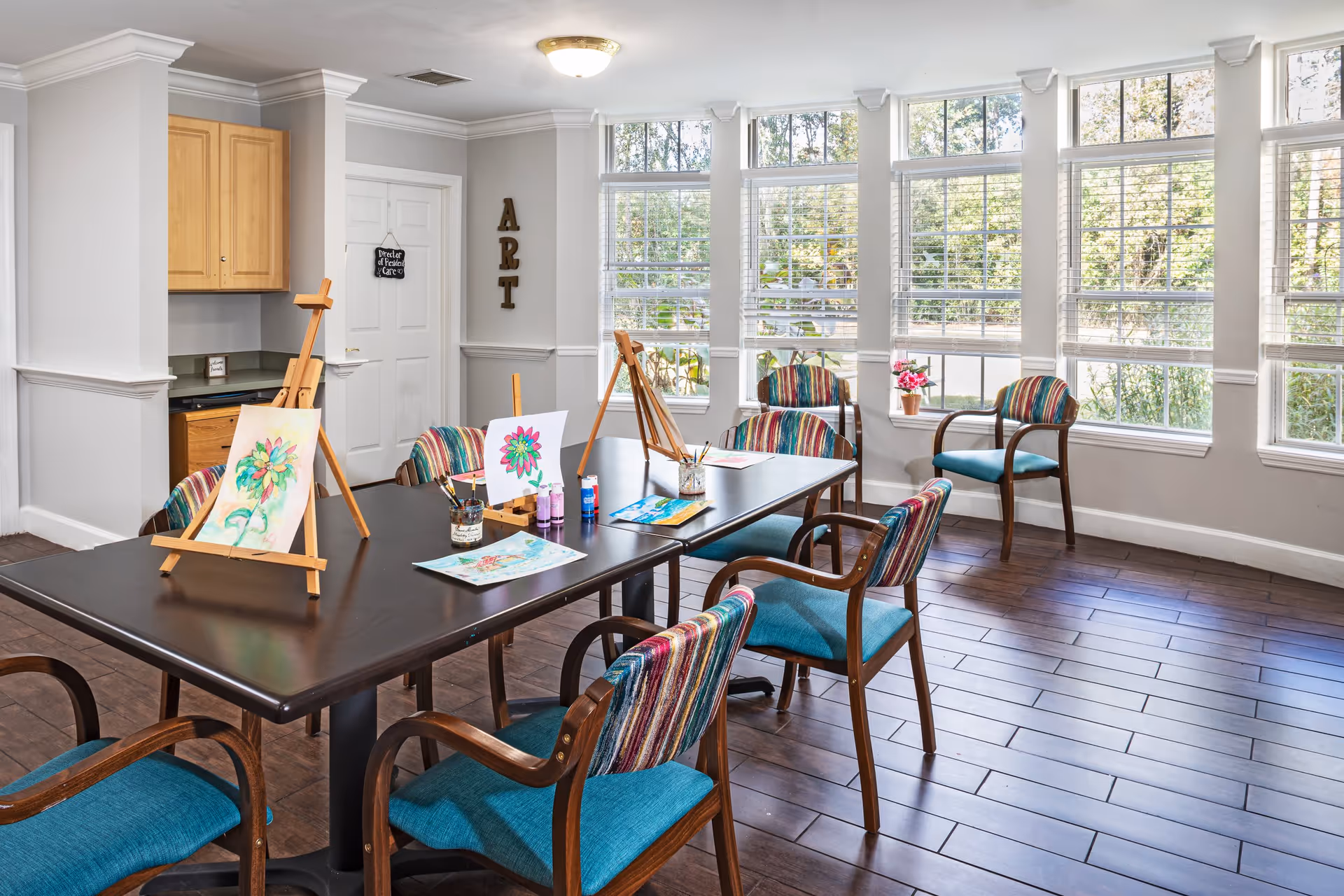 A bright art room with large windows letting in natural light. The room features a dark rectangular table with several colorful chairs around it. On the table are easels holding paintings of flowers, paint bottles, brushes, and paper. The walls are light gray with white trim, and the word 'ART' is displayed vertically on one wall near a white door. The floor is dark wood, and there is a small kitchenette area with wooden cabinets in the corner.