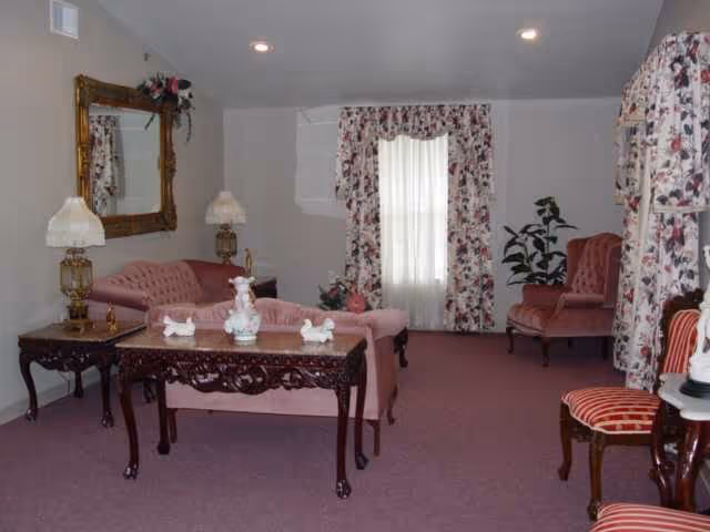 A living room with vintage-style furniture including a pink tufted sofa, matching armchairs, and a wooden coffee table with decorative items. The room has floral curtains on the windows, a large ornate mirror on the wall, two table lamps with white shades, and a potted plant in the corner. The carpet is a muted purple color.