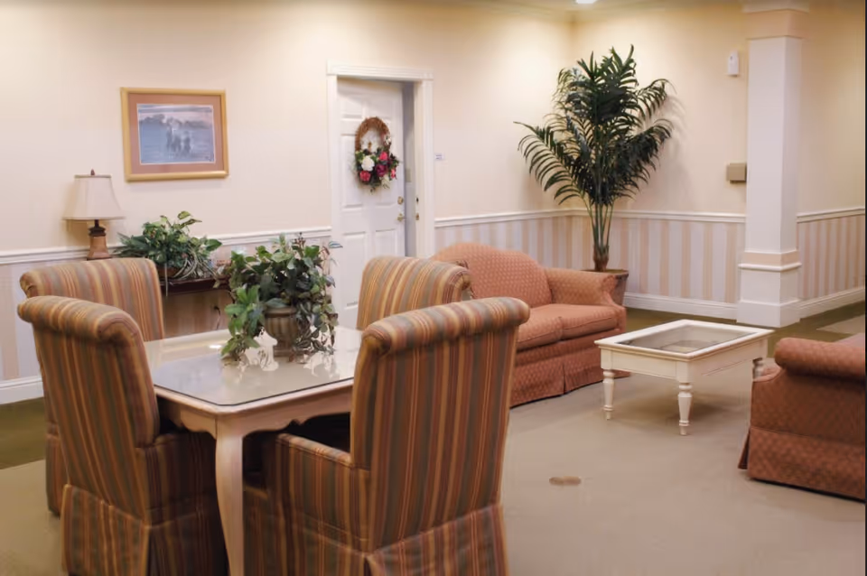 A cozy sitting area in a senior living facility with a small square table surrounded by four striped upholstered chairs. In the background, there is a white door decorated with a floral wreath, a framed picture on the wall, a table with a lamp and plants, a potted tall green plant, and two upholstered sofas with a white coffee table in front of them. The walls are light-colored with a striped wallpaper border and the floor is carpeted.