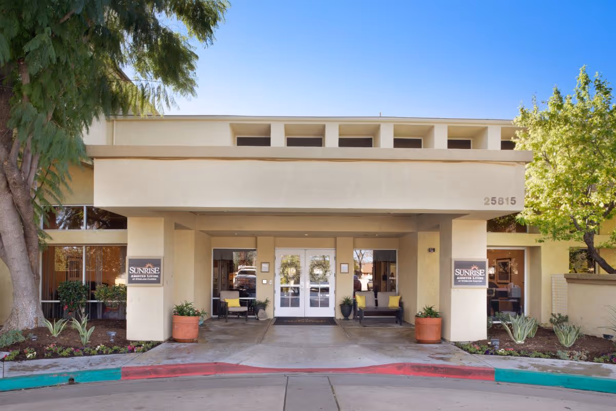 Front entrance of Sunrise At Sterling Canyon showing a covered drive-through entrance with double glass doors, seating, potted plants, and signage.