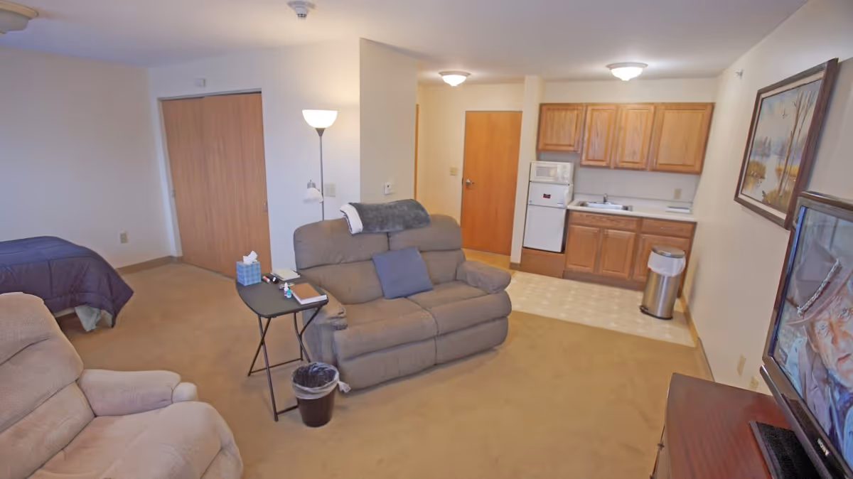A cozy living space in a supportive living community featuring a beige recliner and loveseat with a small black side table holding books and tissues. The room includes a kitchenette with wooden cabinets, a microwave, a mini fridge, and a sink. A bed with a dark comforter is partially visible on the left side. A TV is on the right showing an elderly man, and a framed painting hangs on the wall above the kitchenette. The room has beige carpeting and neutral-colored walls.