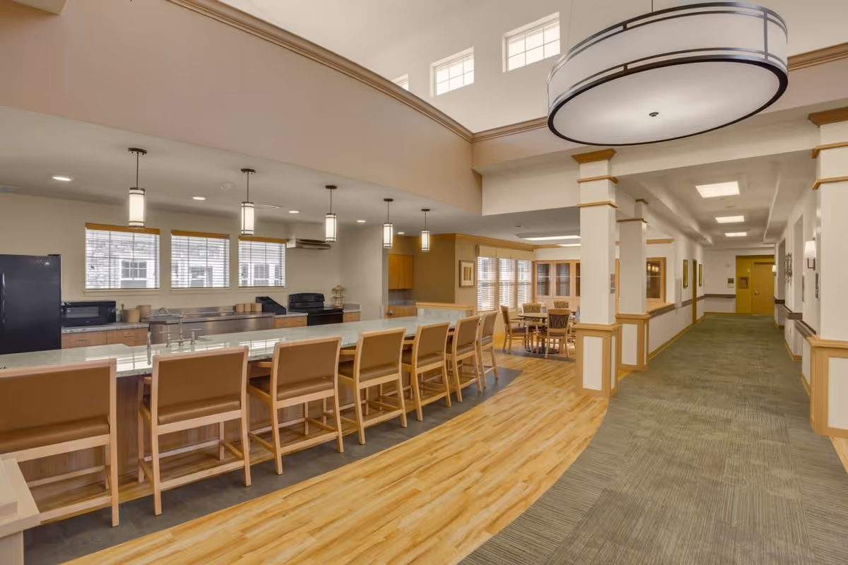 Interior view of a senior living facility kitchen and dining area with a long counter and several chairs, pendant lights hanging from the ceiling, wooden flooring near the kitchen, carpeted hallway, and a round dining table with chairs in the background.