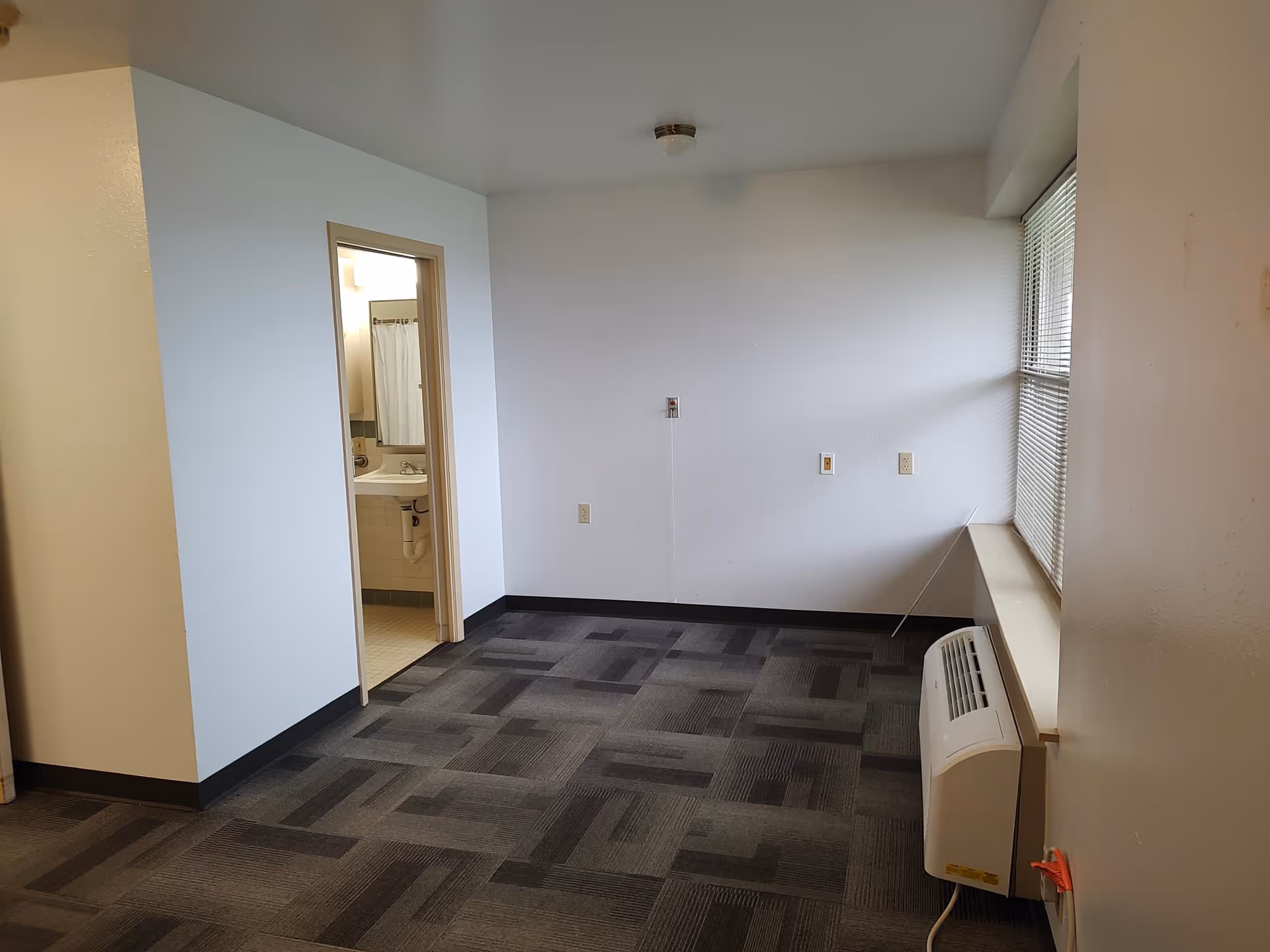 Empty small apartment room with carpeted floor, a window with blinds and a wall-mounted air unit, and an open doorway leading to a bathroom.