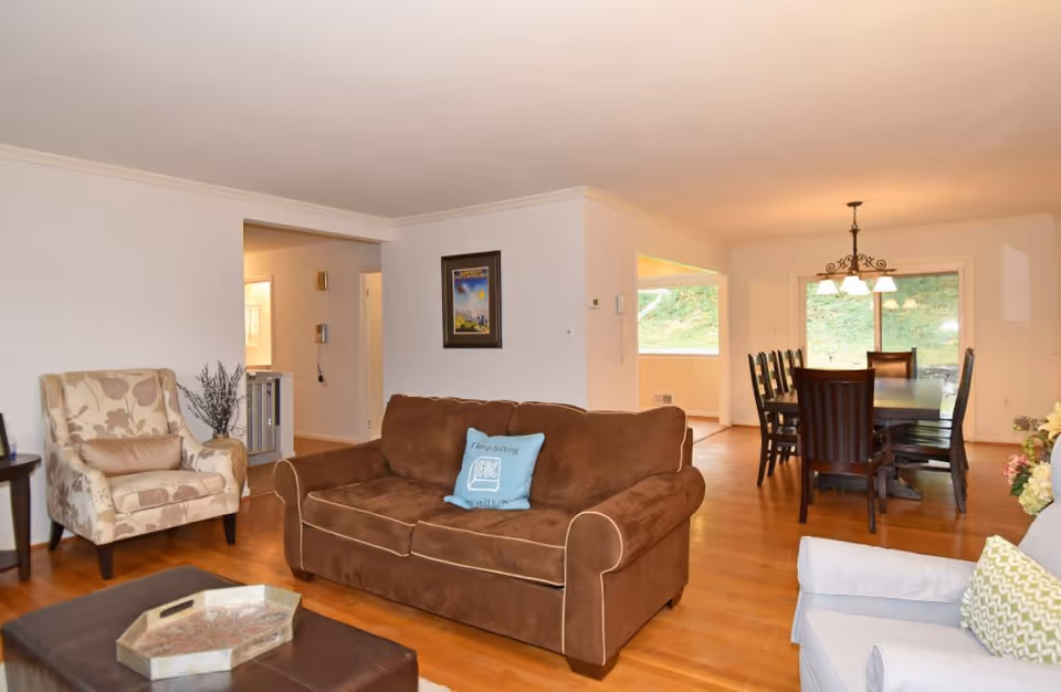 A cozy living room with a brown sofa, a beige patterned armchair, a light gray armchair, and a dark brown ottoman with a decorative tray. The room has wooden flooring and white walls. In the background, there is a dining area with a dark wooden dining table and chairs, a chandelier above the table, and large windows letting in natural light.