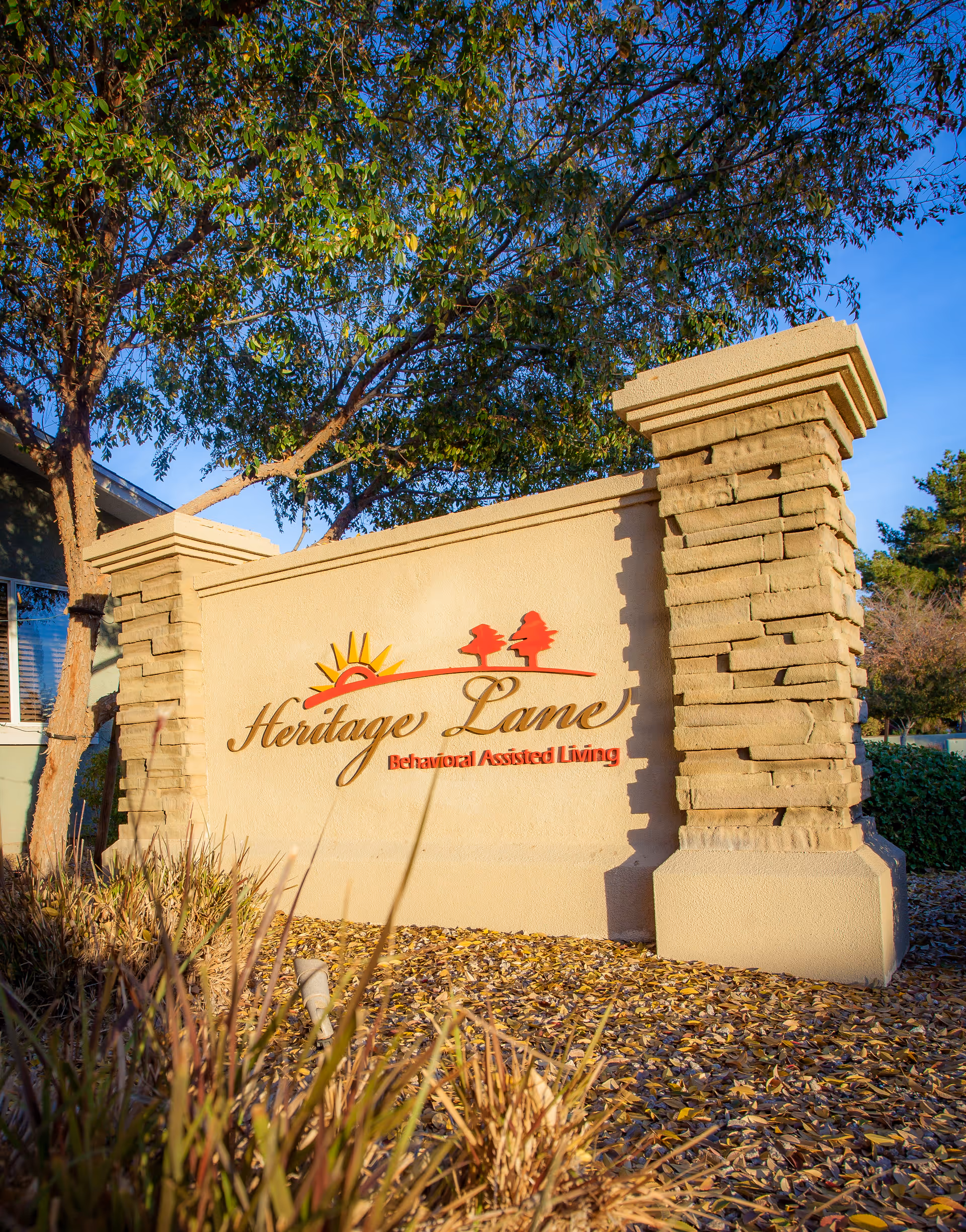 Stone entrance sign that reads 'Heritage Lane Behavioral Assisted Living' set among landscaping and trees.