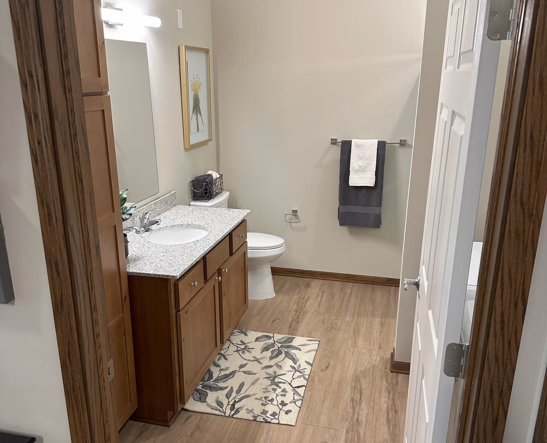 A clean and modern bathroom with wooden cabinetry, a granite countertop with a sink, a toilet, a towel rack with dark and light towels, a floral framed picture on the wall, and a floral patterned rug on the wooden floor.