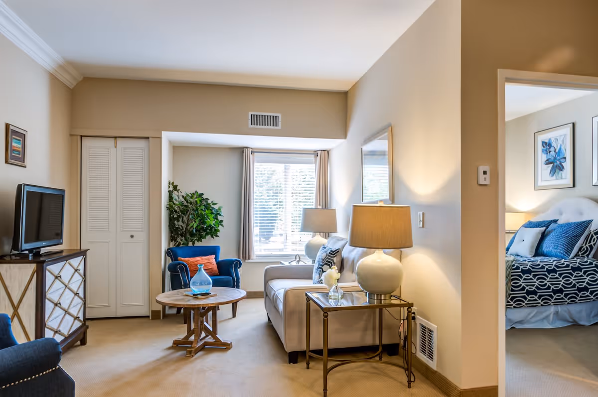 A cozy living room with beige walls and carpet, featuring a beige sofa with patterned pillows, a round wooden coffee table, a blue armchair with an orange pillow, a TV on a wooden cabinet, and a large window with curtains letting in natural light. A doorway leads to a bedroom with a bed dressed in blue and white bedding and floral artwork on the walls.