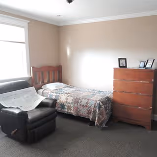 A small bedroom with a single bed covered by a patchwork quilt, a wooden dresser with framed photos on top, a black leather armchair with a newspaper on it, and a window letting in natural light.