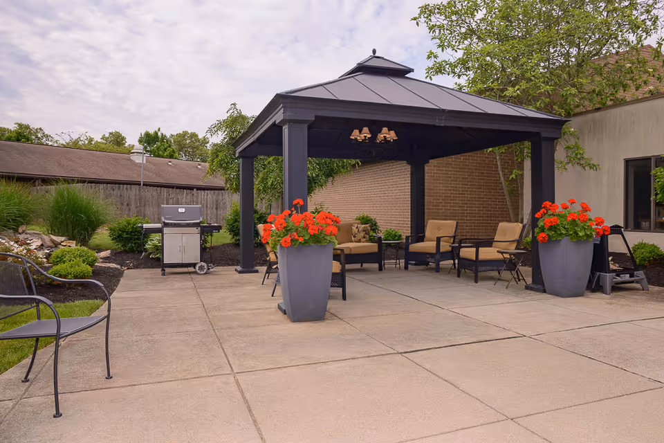 Outdoor patio area at Mayfair Village Retirement Center featuring a black metal gazebo with cushioned chairs and a small table underneath. Two large planters with red flowers flank the entrance to the gazebo. There is a barbecue grill to the left and some greenery and bushes along the fence in the background.