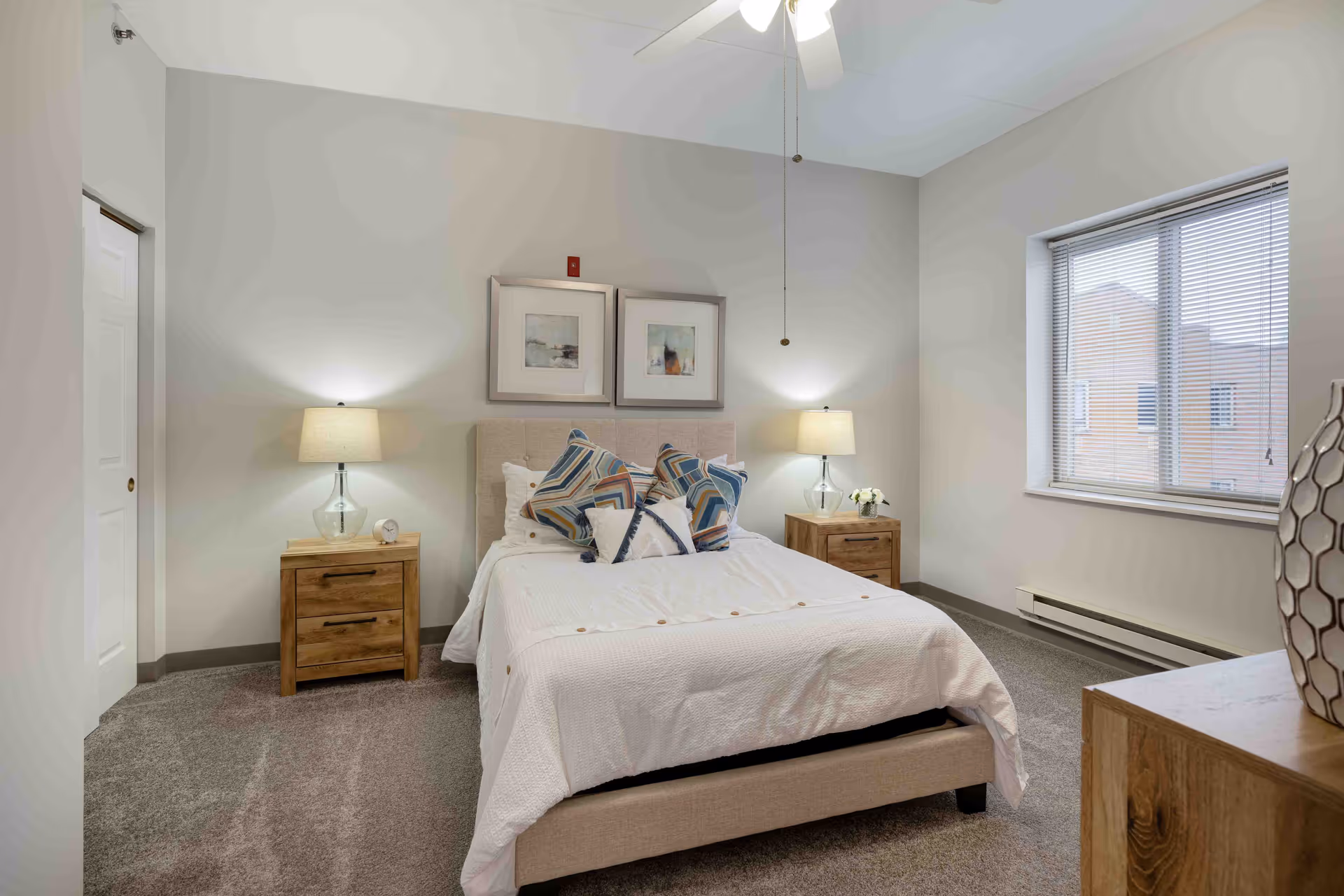 A neatly arranged bedroom with a bed in the center, adorned with multiple decorative pillows and a white bedspread. On each side of the bed are wooden nightstands with glass lamps and small decorative items. Two framed artworks hang above the bed. A window with blinds is on the right wall, and a ceiling fan with a light is visible above. The room has carpeted flooring and neutral-colored walls.