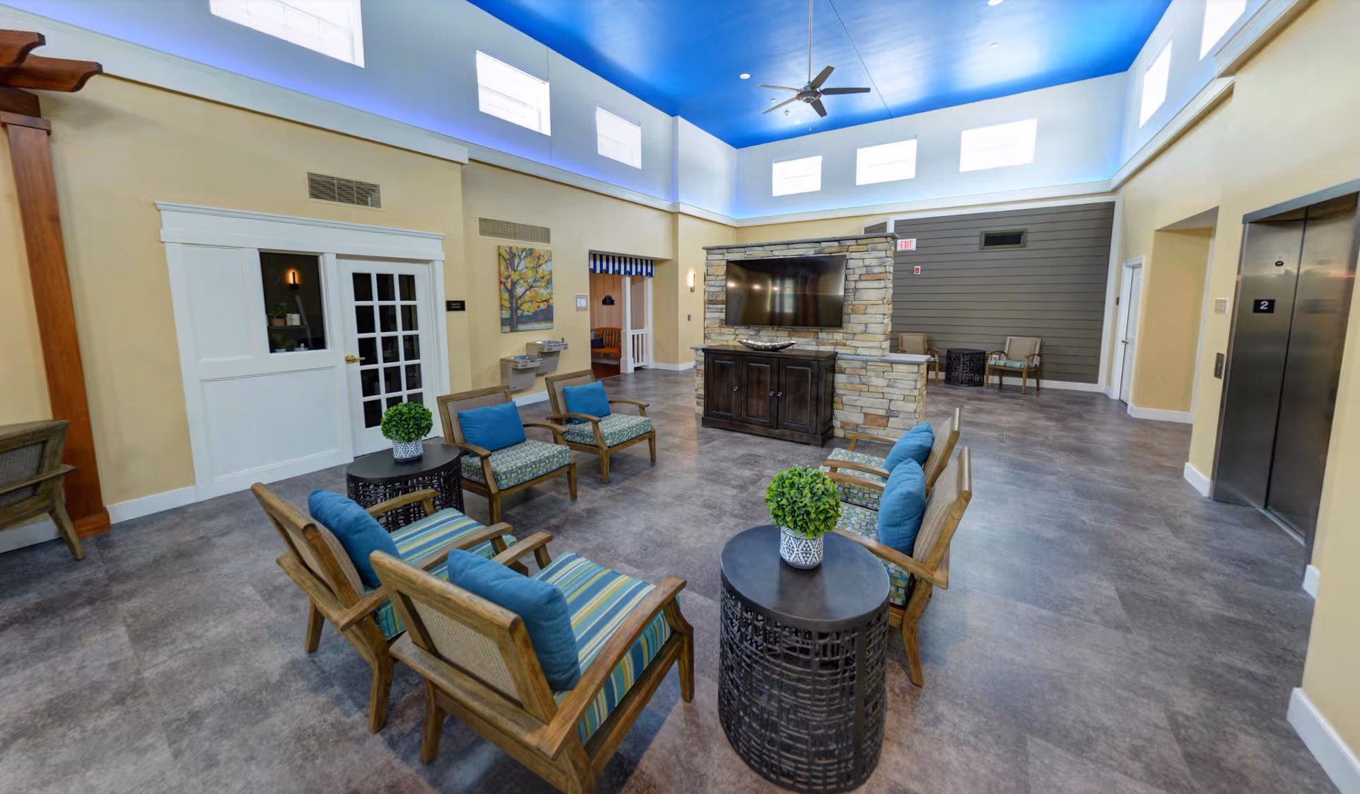 A spacious senior living facility common area with a high blue ceiling and multiple small windows near the ceiling. The room features several wooden chairs with blue and striped cushions arranged around small round tables with potted plants. There is a large flat-screen TV mounted on a stone wall divider, and an elevator is visible on the right side. The walls are painted beige with white trim, and there is a ceiling fan above.