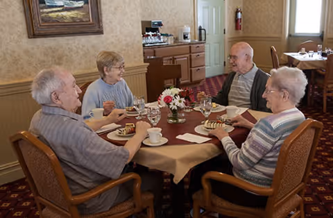 Four elderly residents sit around a table in a dining room enjoying coffee and dessert.