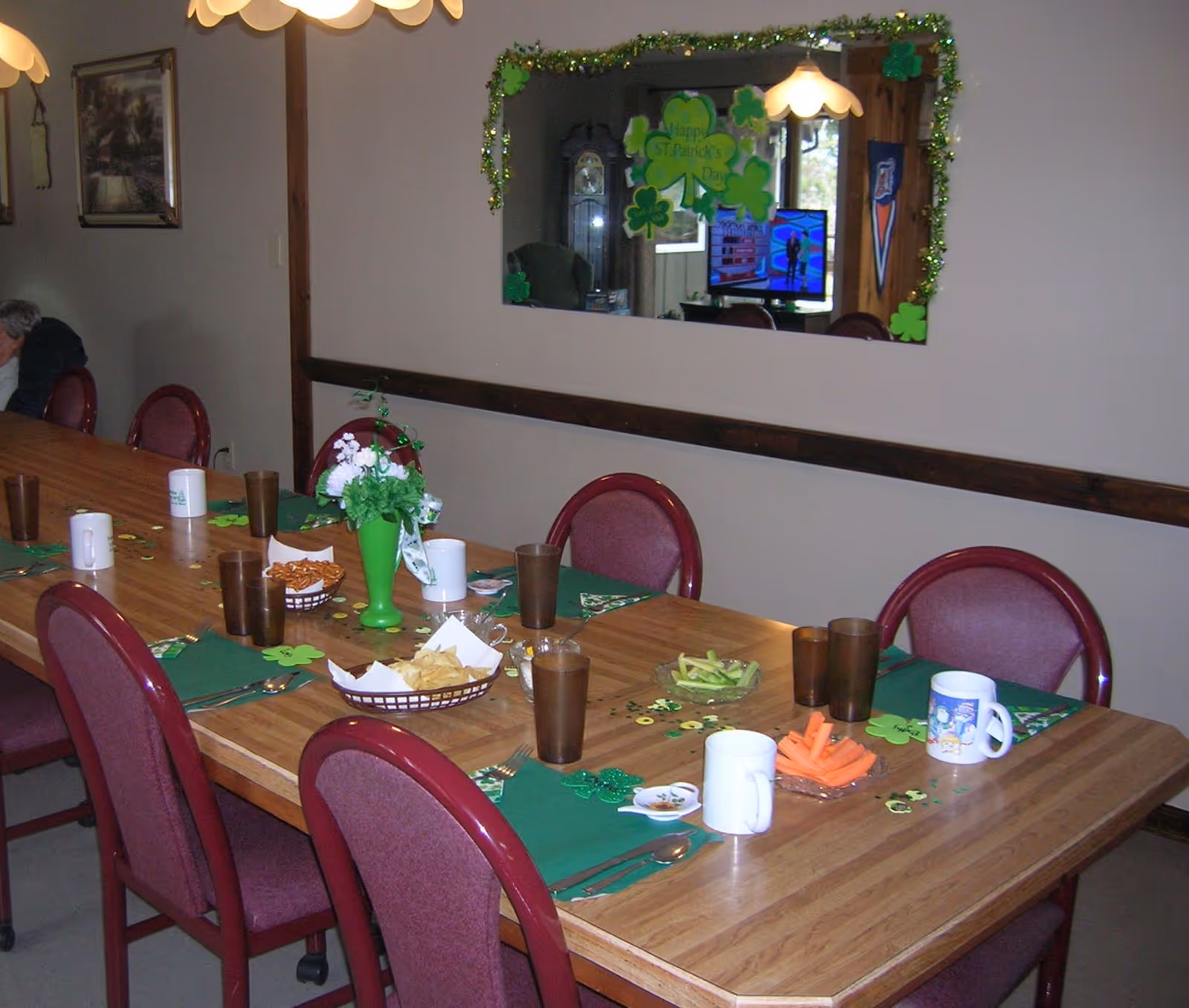 A dining room table set with green placemats, brown drinking glasses, white mugs, and plates of snacks including pretzels, chips, celery, and carrot sticks. The table is decorated with St. Patrick's Day themed items such as shamrocks and a green vase with flowers. A large mirror on the wall reflects a television and additional decorations. Several maroon chairs surround the table.