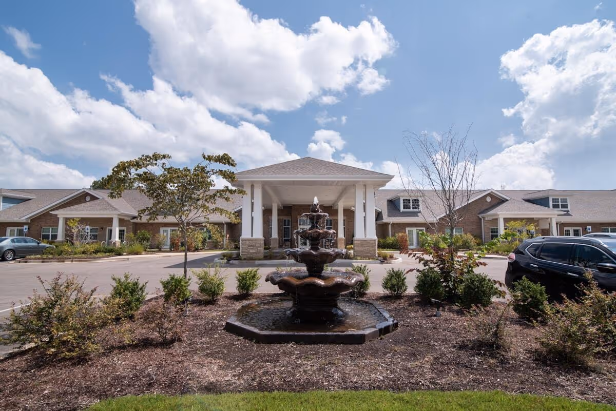 Front exterior view of a single-story senior living facility building with a covered entrance supported by white columns. In front of the building is a multi-tiered water fountain surrounded by landscaped bushes and small trees. Cars are parked on either side of the entrance driveway under a partly cloudy sky.