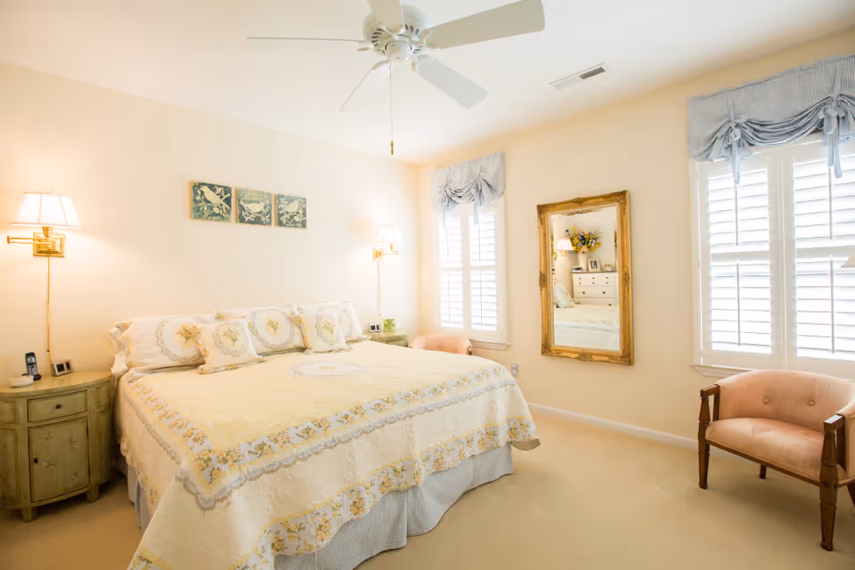 A bright and cozy bedroom with a large bed covered in a floral quilt and matching pillows. The room features two wall-mounted lamps on either side of the bed, a ceiling fan, two windows with white shutters and blue valances, a vintage-style wooden chair with pink upholstery, and a large ornate gold-framed mirror reflecting a dresser with flowers and picture frames.