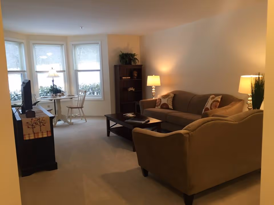 Cozy furnished living room with a sofa, armchair, coffee table, TV and a small dining nook by bay windows.