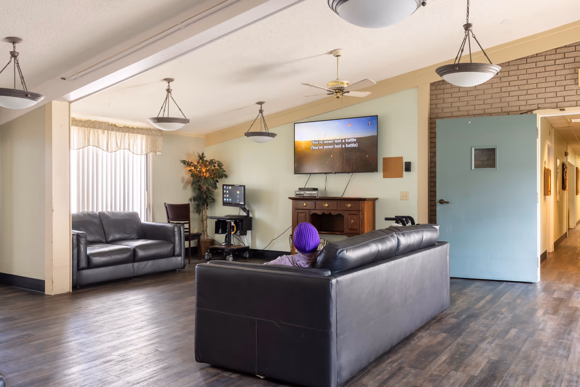 A senior living facility common area with two black leather sofas, a person sitting on one sofa watching a wall-mounted TV displaying karaoke lyrics, a computer on a small desk, a potted plant, and a blue door leading to a hallway.