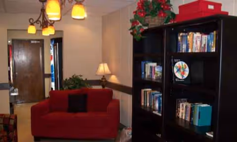 Red sofa beside a tall bookshelf in a cozy lobby with a lamp and plants.