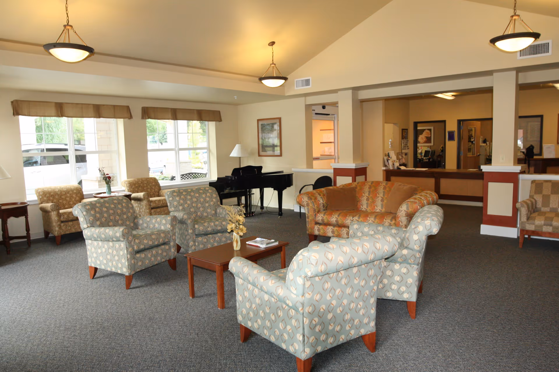 A spacious senior living facility common area with patterned armchairs and a sofa arranged around a wooden coffee table. Large windows let in natural light, and a black grand piano is positioned near the wall. The area has beige walls, carpeted floor, and ceiling lights. In the background, there is a reception desk with office doors behind it.