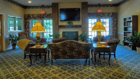 Comfortable common lounge with patterned sofas and chairs facing a brick fireplace and wall-mounted TV, flanked by table lamps, windows, and bookshelves.