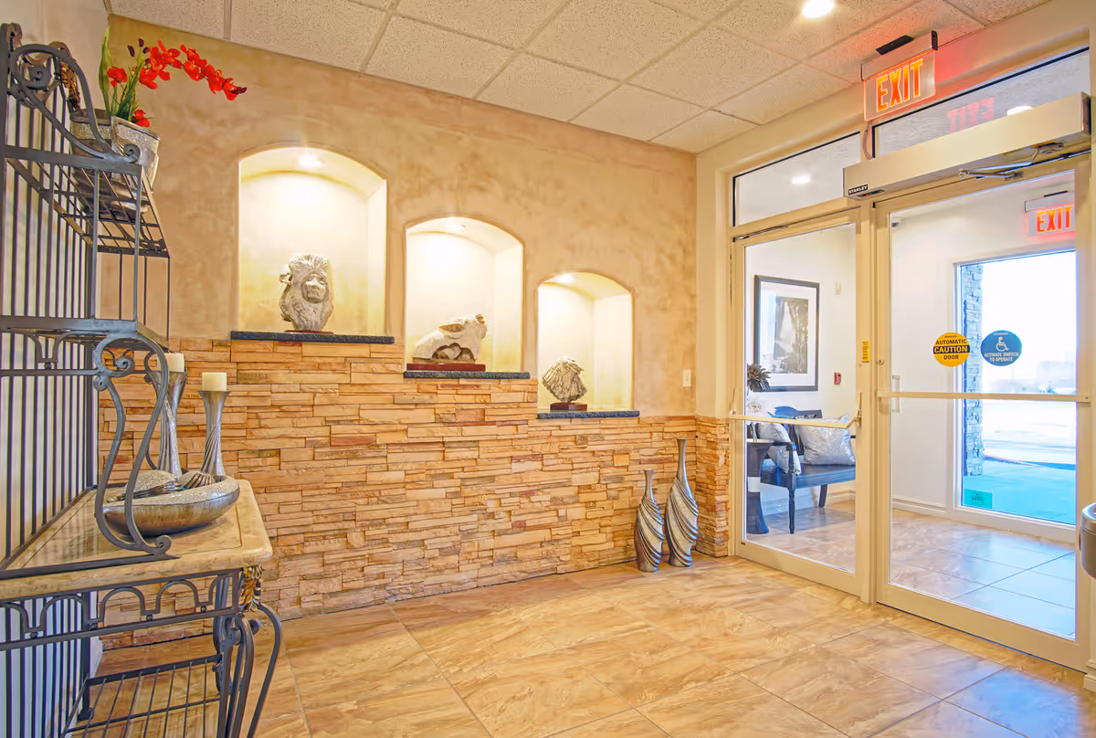 Interior view of an assisted living facility entrance area with a decorative stone half-wall featuring three illuminated niches displaying sculptures. To the left, there is a metal and glass shelving unit with decorative items. On the right, glass double doors with exit signs lead outside, showing a glimpse of a seating area with a framed picture on the wall.