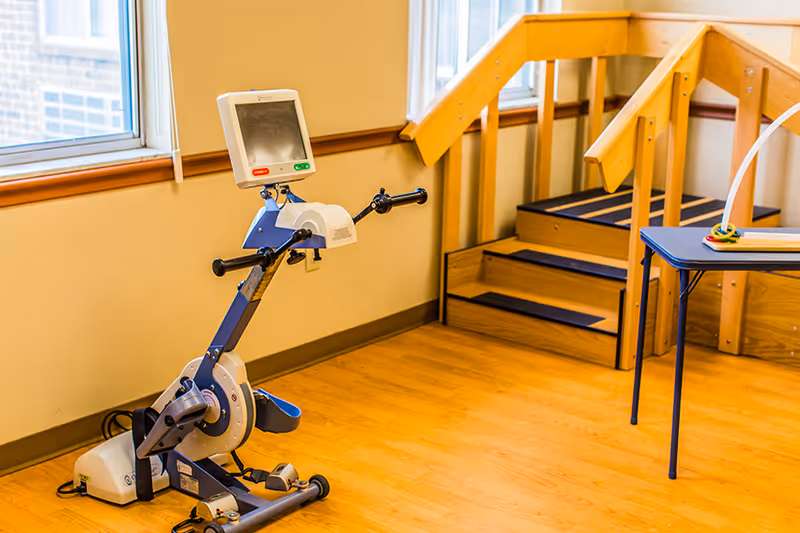 A small exercise bike with a digital screen is positioned near a window in a room with wooden flooring. Next to it is a wooden staircase with handrails, and a table with a toy on top is partially visible on the right side.