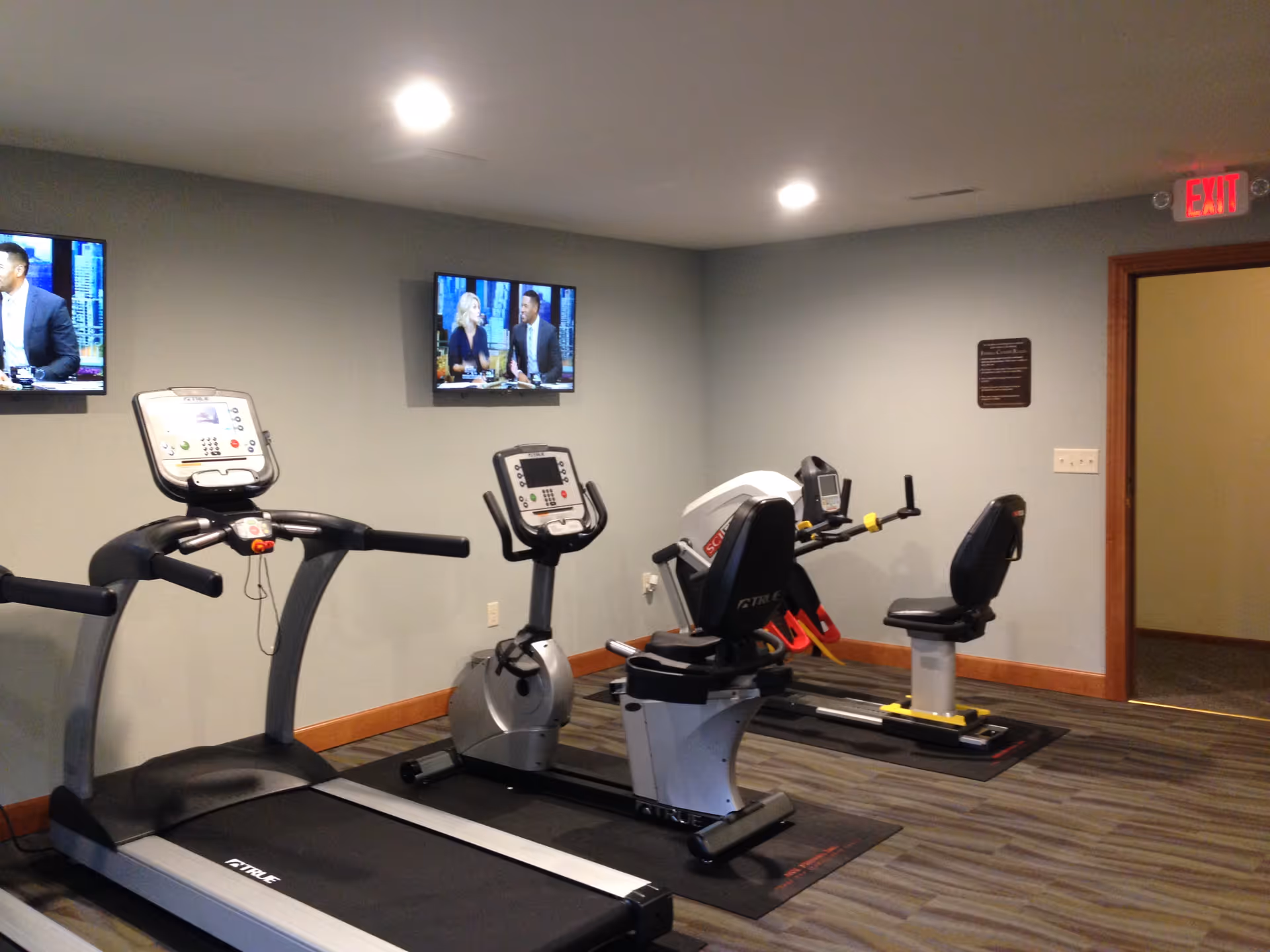 Indoor fitness room with exercise equipment including a treadmill, an upright stationary bike, and a recumbent bike. Two wall-mounted televisions are visible on the wall, and there is an exit door with a red exit sign above it.
