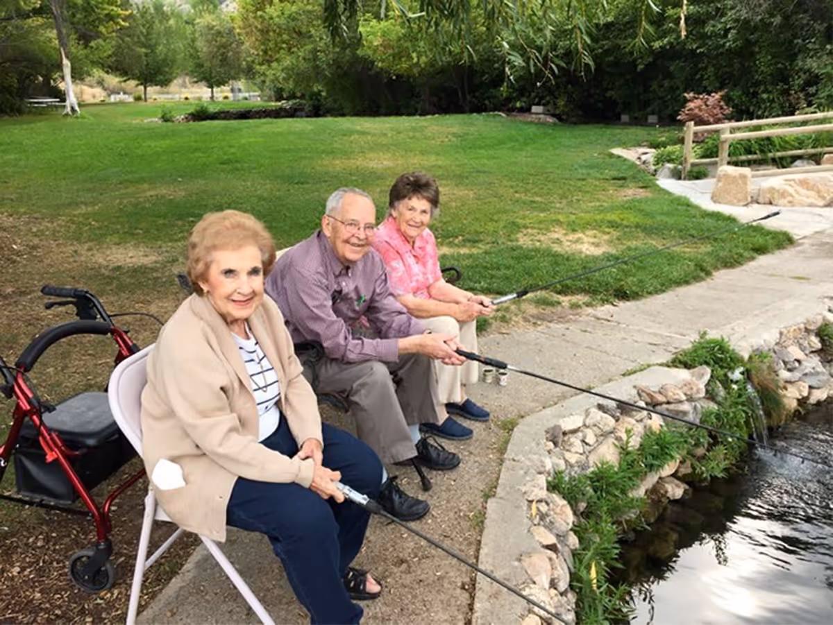 Three elderly people sitting on chairs by a small pond, fishing with fishing rods. They are outdoors on a grassy area with trees and bushes in the background. One woman has a red walker beside her.