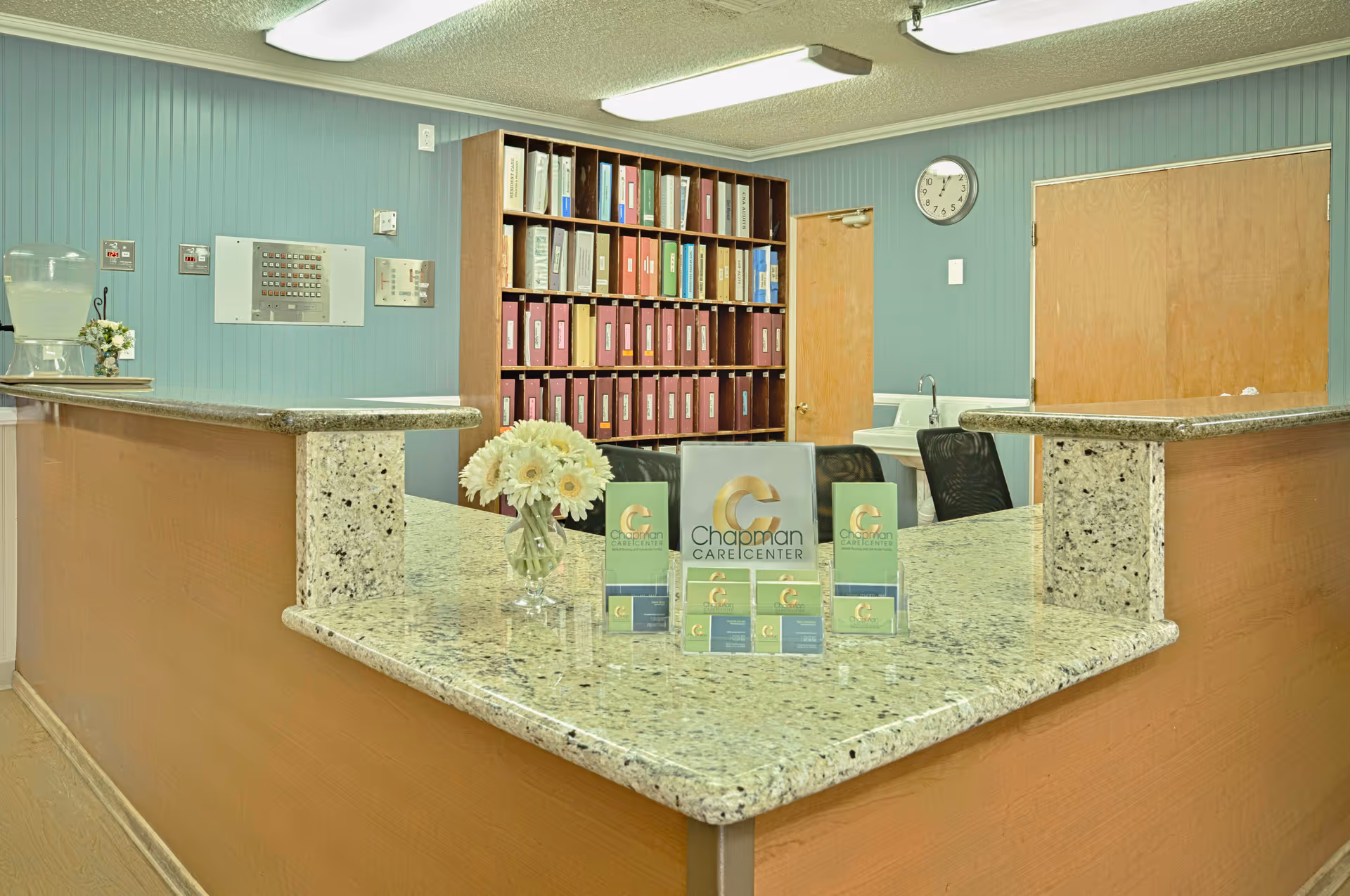 Reception desk area at Chapman Care Center with a granite countertop, a vase of white flowers, informational brochures, a water dispenser on the side, a bookshelf filled with binders, a clock on the wall, and two wooden doors in the background.