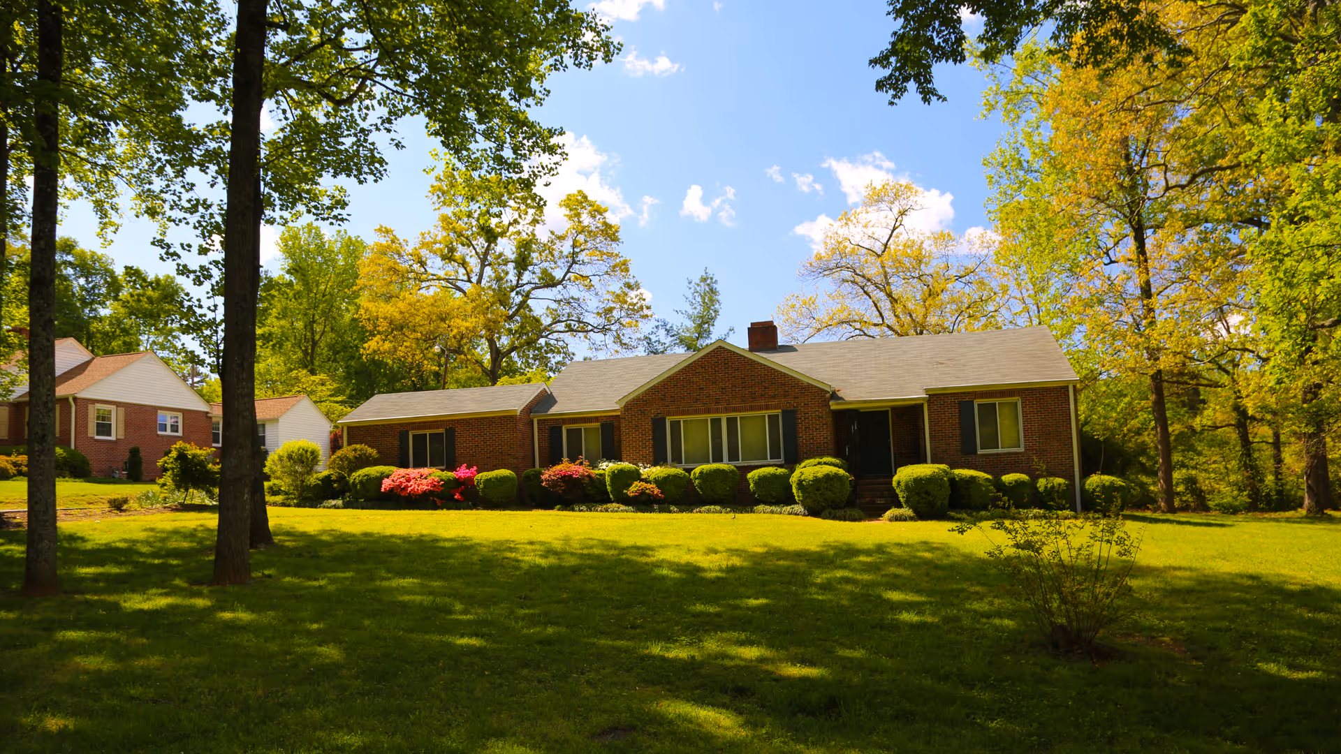 A single-story brick house with a gray roof surrounded by green grass, bushes, and trees under a blue sky with some clouds.