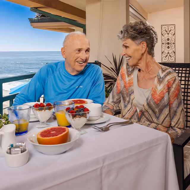 An elderly man and woman sitting at a table outdoors, enjoying a meal with bowls of fruit, glasses of orange juice, and a halved grapefruit on the table. They are smiling and looking at each other with the ocean visible in the background.