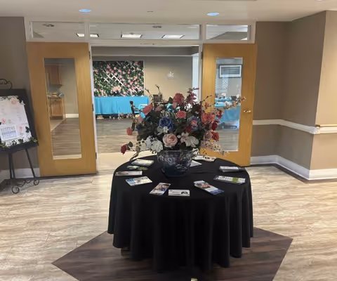 A round table covered with a black tablecloth is centered in a room with light-colored tiled flooring. On the table is a large floral arrangement with various colorful flowers in a decorative vase, surrounded by brochures. Behind the table are double wooden doors with glass panels, leading to a room with tables covered in blue tablecloths and a floral wall decoration.