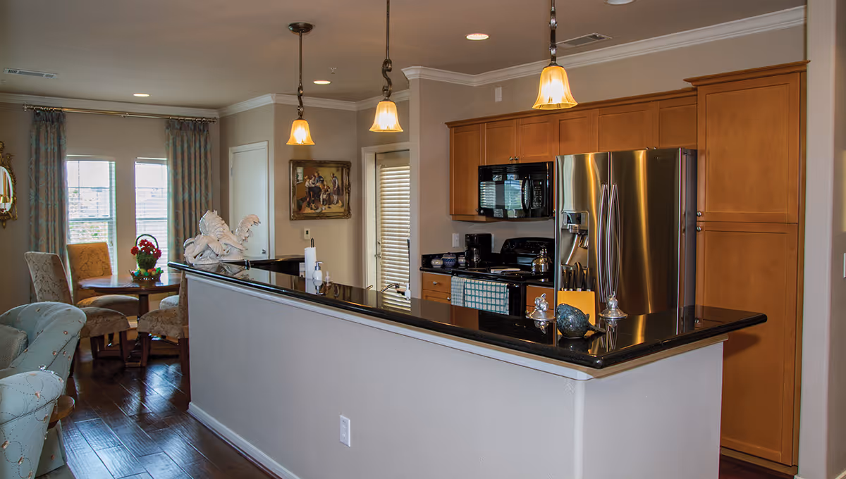 Interior view of a modern kitchen with wooden cabinets, stainless steel refrigerator, black countertop, and three hanging pendant lights. In the background, there is a small dining area with a round table, four upholstered chairs, and a window with curtains.