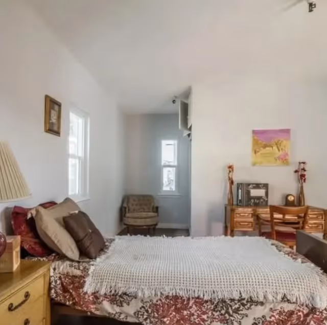 A bedroom with a bed covered in a white textured blanket and floral bedspread, several pillows, a wooden nightstand with a lamp, a wooden desk with a chair, decorative items, and a painting on the wall. There is a window on the left wall and a small armchair in a nook near the back wall.