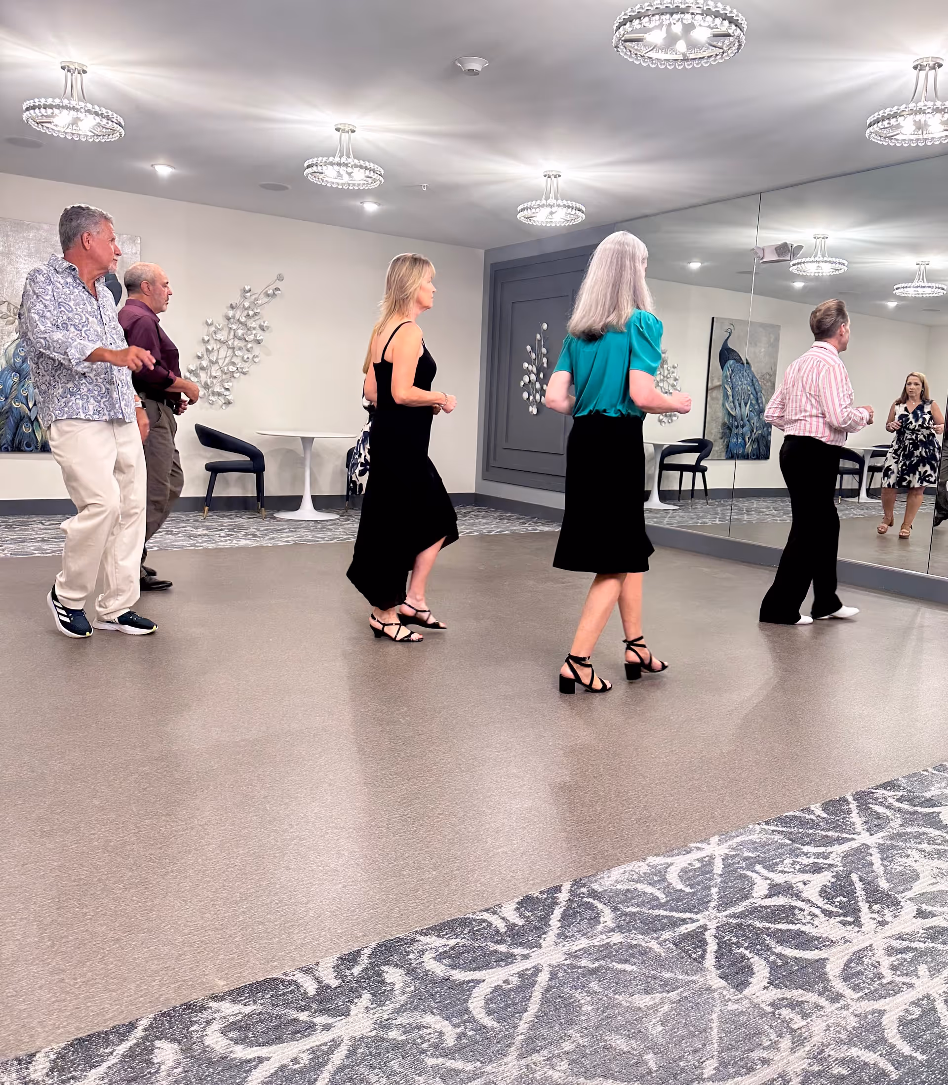 A group of five adults standing in a spacious room with a large wall mirror, practicing dance steps. The room has modern light fixtures on the ceiling, patterned carpet along the edges, and decorative wall art including a peacock painting and metal wall sculptures. There are a couple of small tables and chairs against the walls.