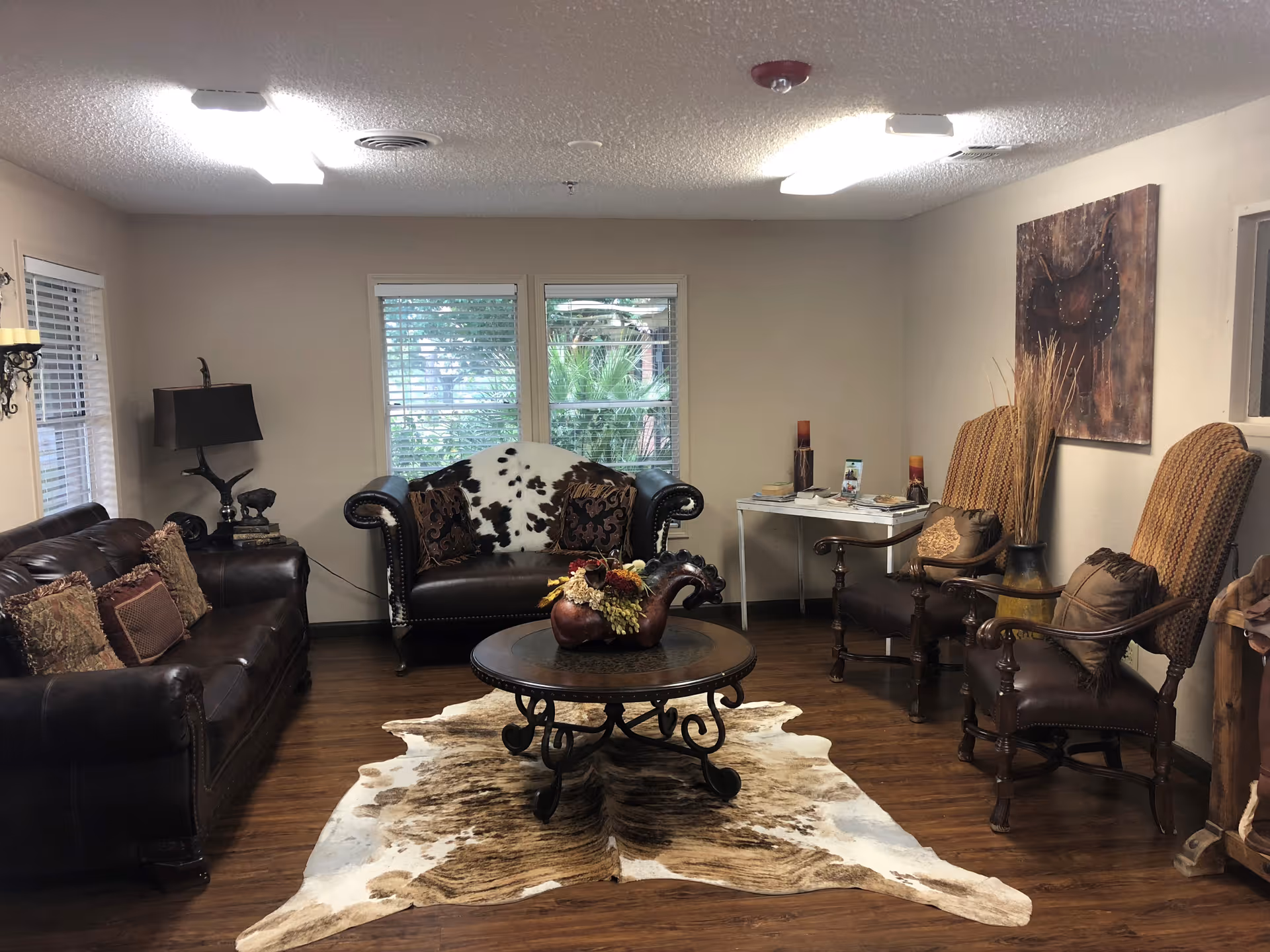 A cozy living room with dark leather sofas and armchairs arranged around a round wooden coffee table. The floor has a cowhide rug, and there are decorative pillows on the seating. Two windows with blinds allow natural light, and a painting and decorative items are on the walls and tables.