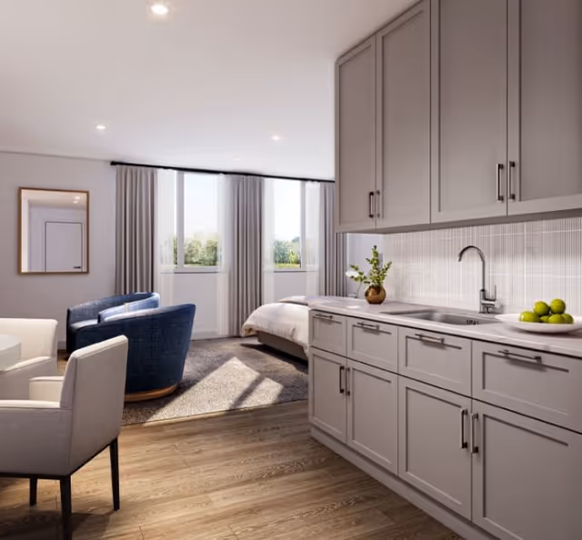 A bright and modern senior living studio apartment featuring a kitchenette with gray cabinets, a sink, and a countertop with a vase and a bowl of green apples. The room includes a bed near large windows with curtains, a blue armchair, a beige armchair, and a wall mirror reflecting a door.