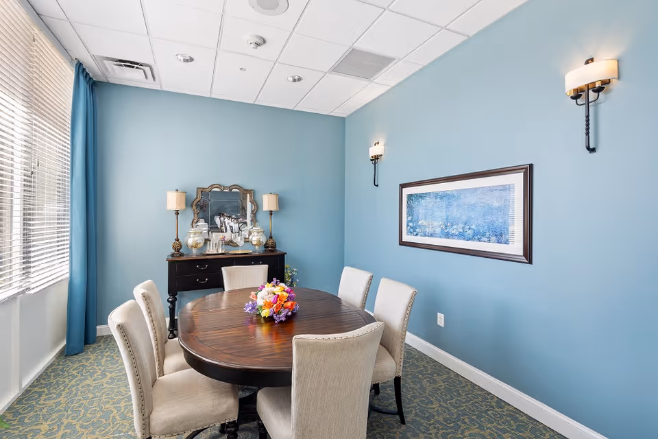 A small dining room with a round wooden table surrounded by six beige upholstered chairs. A colorful flower arrangement is placed in the center of the table. The walls are painted light blue, with two wall sconces and a framed artwork on one wall. A black sideboard with a decorative mirror and two table lamps is positioned against another wall. Large windows with blinds and blue curtains allow natural light into the room.