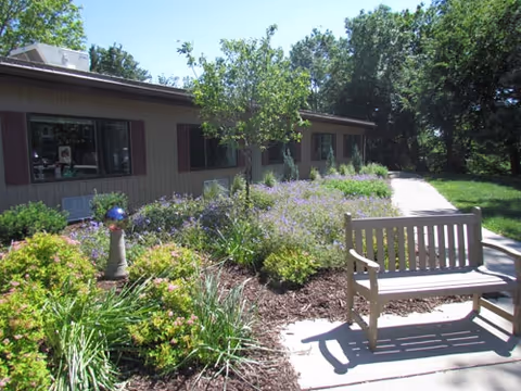 Outdoor garden area with a wooden bench on a paved path, surrounded by various green plants, bushes, and a small tree. A single-story building with windows and brown shutters is visible in the background under a clear blue sky.