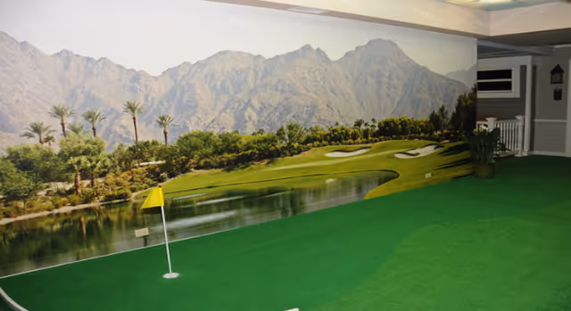 Indoor putting green area with artificial grass and a small yellow flag on a putting hole. The back wall features a large mural of a golf course with a pond, palm trees, and mountains in the background. There are some plants and a small porch area on the right side of the room.