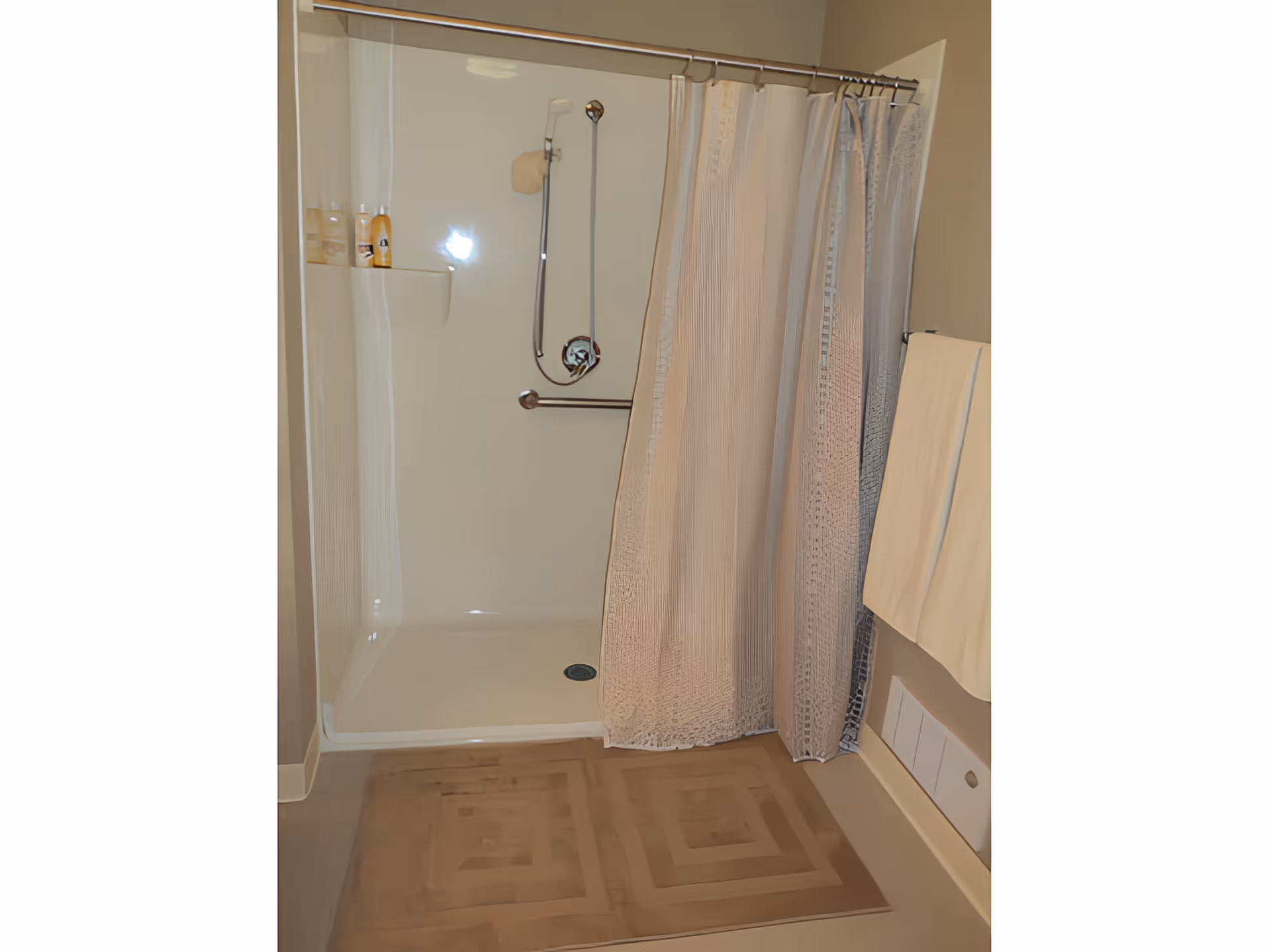 Shower stall with a curtain, grab bar, handheld shower head, towel on a rack and a bath mat on the floor.