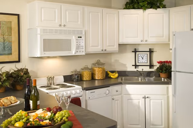 White kitchenette with upper cabinets, microwave, stove, sink and refrigerator, and a countertop display of fruit, wine and jars.