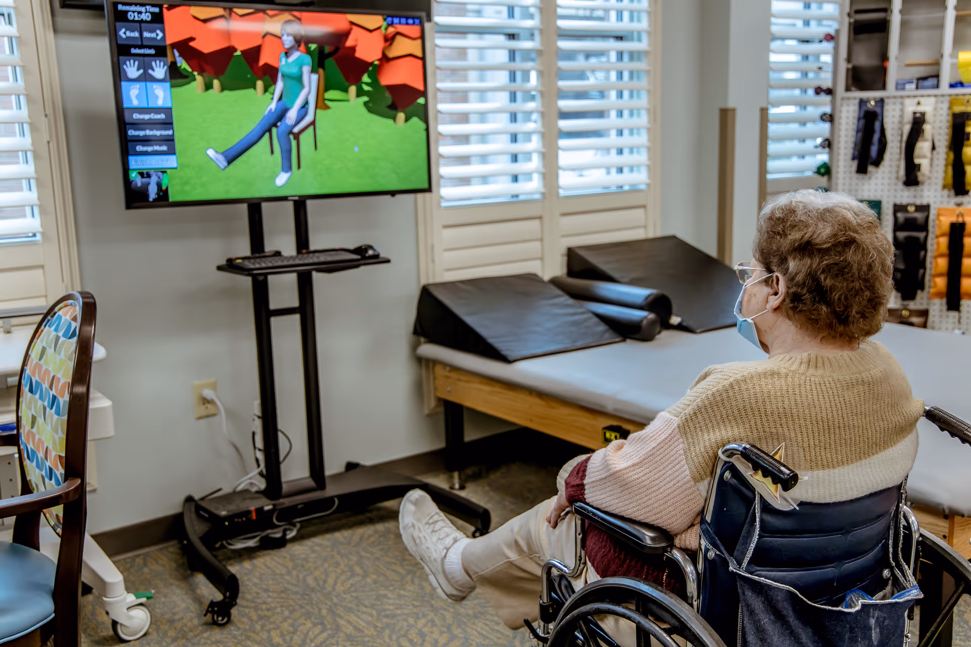 An elderly person in a wheelchair wearing a face mask is seated in a room with medical equipment and therapy mats. The person is watching a large screen displaying a virtual exercise program featuring an animated figure performing leg stretches. The room has windows with white shutters and various therapy supplies on the walls.