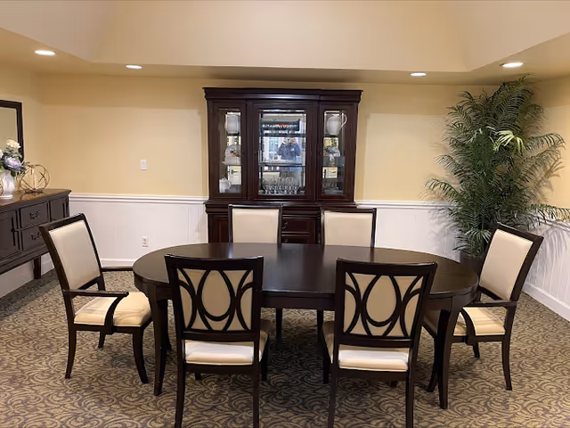 A dining room with a dark wooden oval table surrounded by six cushioned chairs with decorative backs. Behind the table is a dark wooden china cabinet with glass doors displaying glassware and decorative items. To the left is a sideboard with a floral arrangement and decorative objects. A large green potted plant is placed in the right corner. The walls are painted light yellow with white wainscoting, and the floor is carpeted with a patterned design.