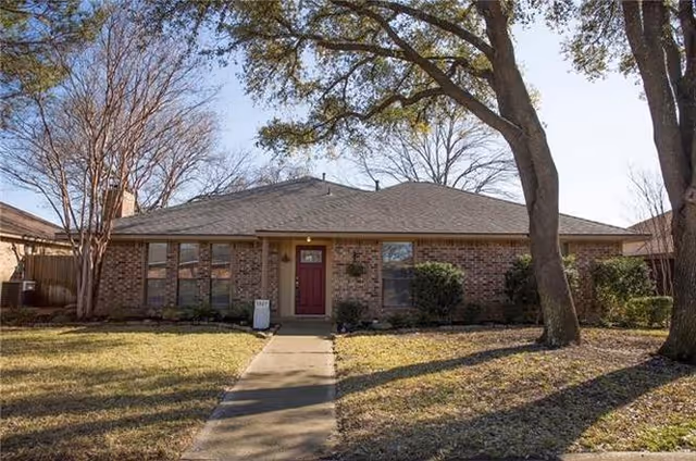 Single-story brick residential building with a gray shingled roof, a red front door, and several windows. There is a concrete walkway leading to the entrance, with trees and shrubs in the front yard under a clear sky.