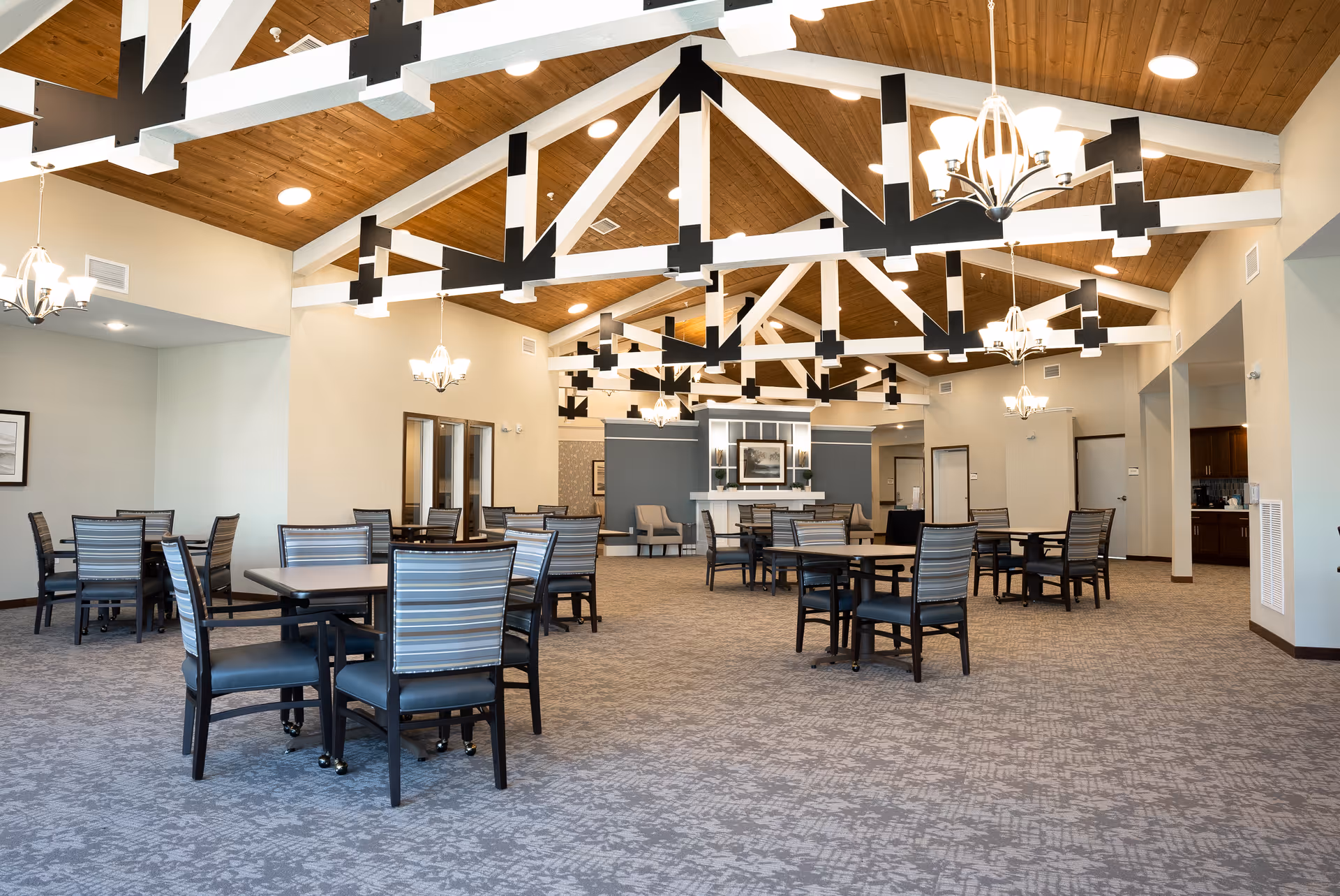 Spacious dining room with multiple tables and chairs arranged on a carpeted floor. The ceiling features exposed wooden beams with black accents and several hanging chandeliers providing warm lighting. The walls are light-colored, and there is a fireplace with framed artwork above it in the background.