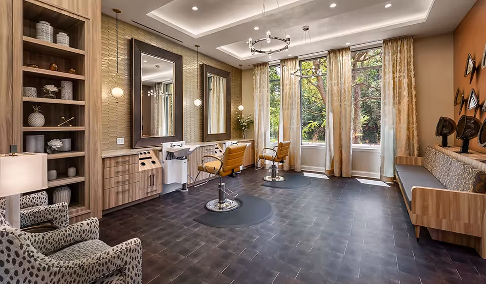 Bright salon-style interior with styling chairs, large mirrors, a hair-washing sink, seating area, and tall windows overlooking trees.