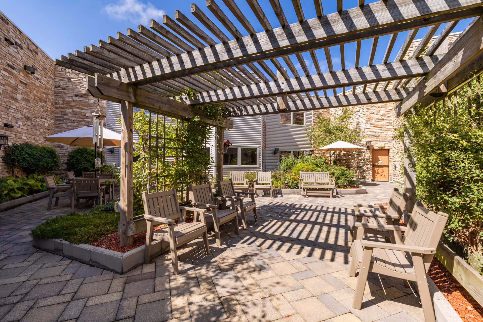 Outdoor courtyard area with wooden pergola casting shadows on the paved ground. Several beige chairs are arranged under the pergola and around the courtyard. There are green plants and bushes along the edges, stone and siding walls of the building surround the space, and two umbrellas provide shade for additional seating areas.